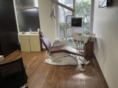 An empty dental treatment room with a reclined chair, overhead light, and a window looking out to greenery.
