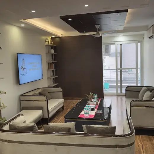 A waiting lounge with grey sofas, a coffee table, shelving with plants and books, and a wall-mounted TV.